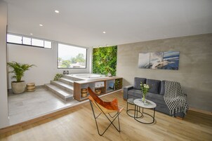 Una sala de estar moderna con un sofá, una mesa de centro, una silla, un ventanal grande, una zona de cocina con un lavamanos y una pared verde.