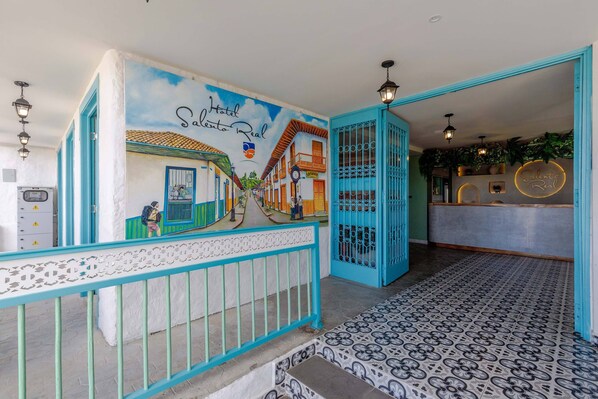 Un vestíbulo de hotel con un mural que representa una escena callejera, una recepción y una escalera con azulejos decorativos.