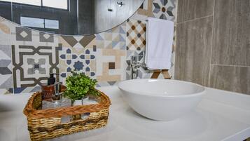 Un bagno con un ampio lavandino bianco, uno specchio, una parete rivestita di piastrelle decorate e un cesto di vimini con articoli da toilette.