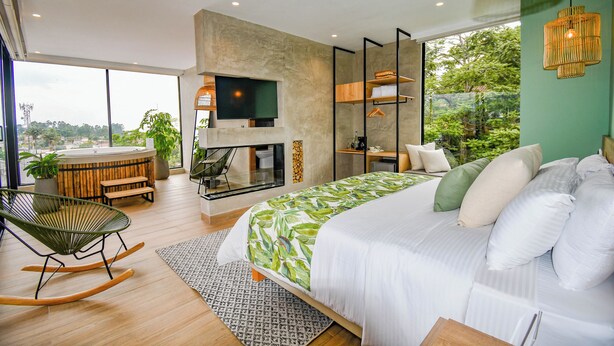 A modern bedroom with a large bed, a TV, a seating area with a chair and table, and a view of greenery outside.