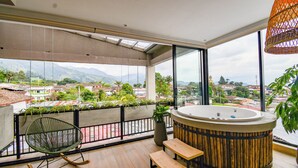 A balcony with a wooden floor, a rocking chair, a bench, a hot tub, and a view of the city and mountains.