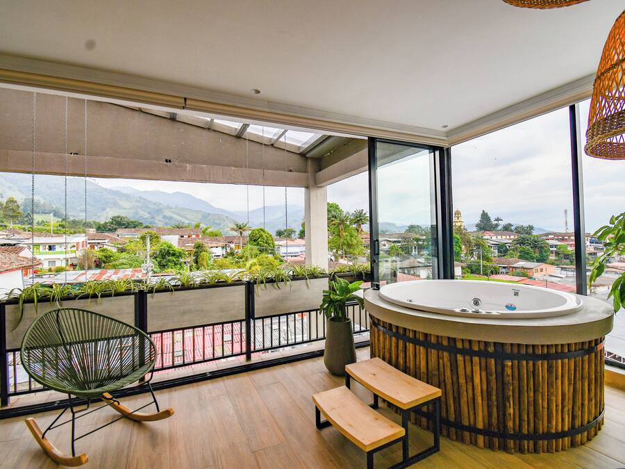 A balcony with a wooden floor, a rocking chair, a bench, a hot tub, and a view of the city and mountains.