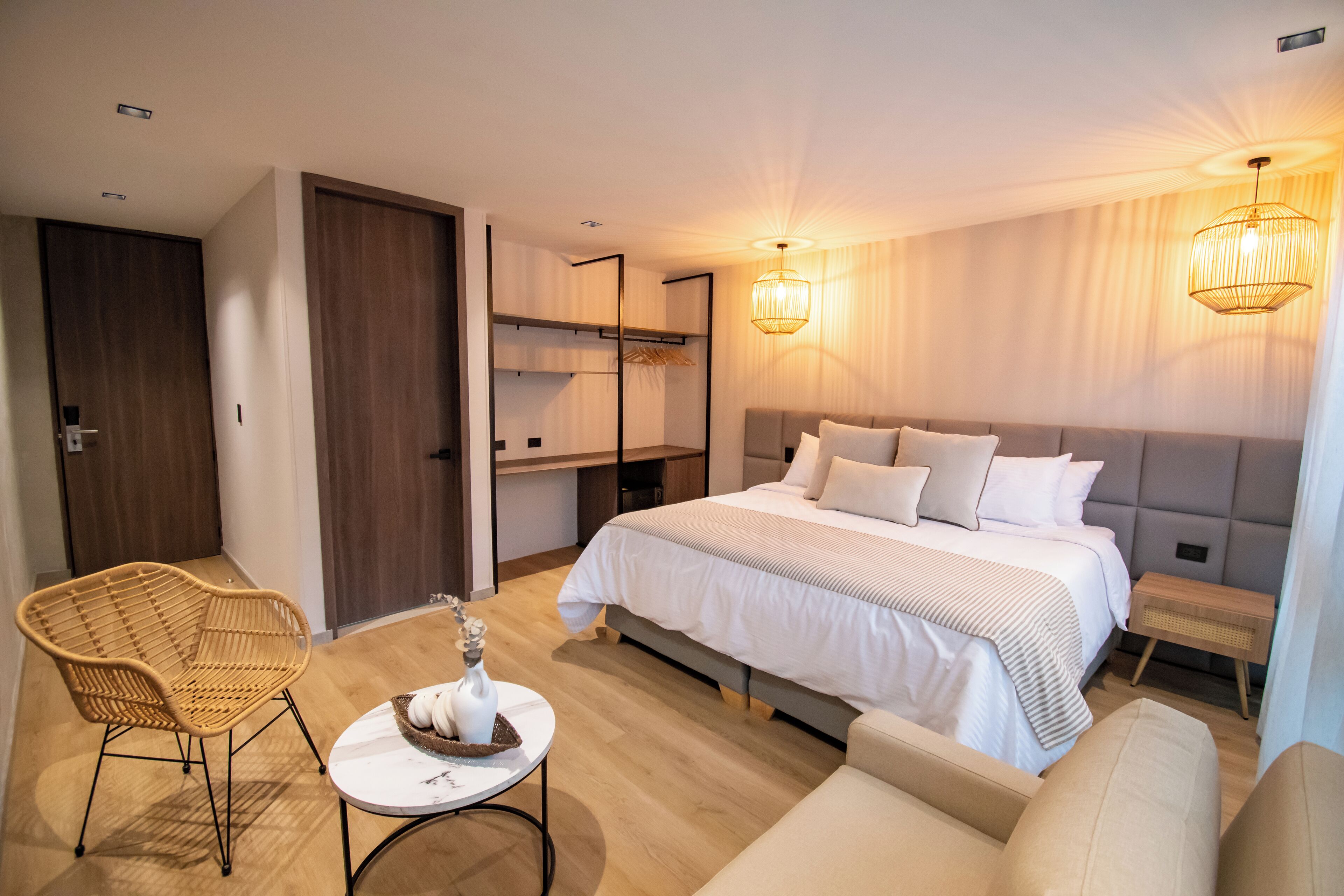 A modern hotel room with a large bed, a sofa, a wicker chair, and a small table with decorative items.