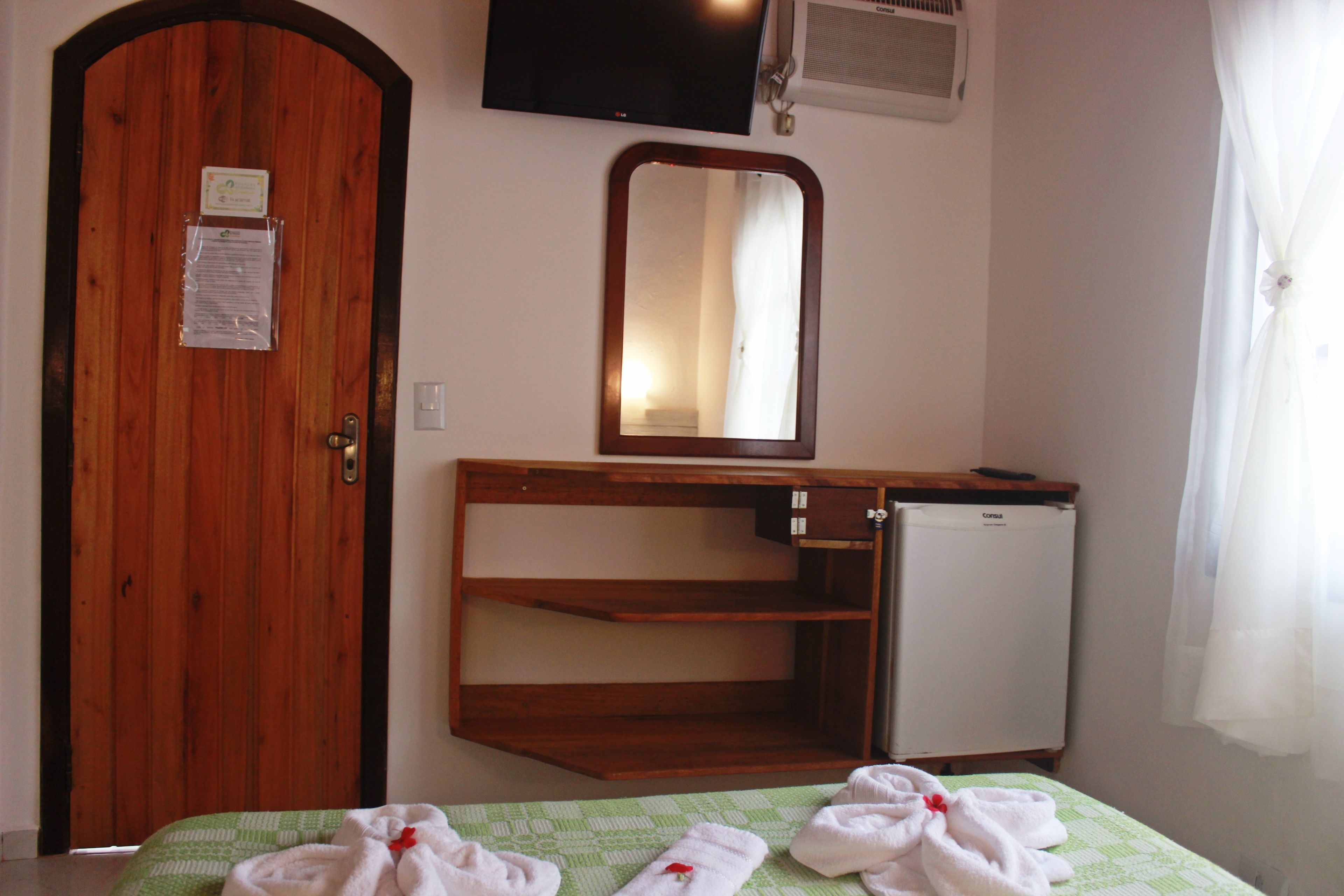 A room with a wooden door, a small refrigerator, a mirror, and a bed with green bedding and towels.