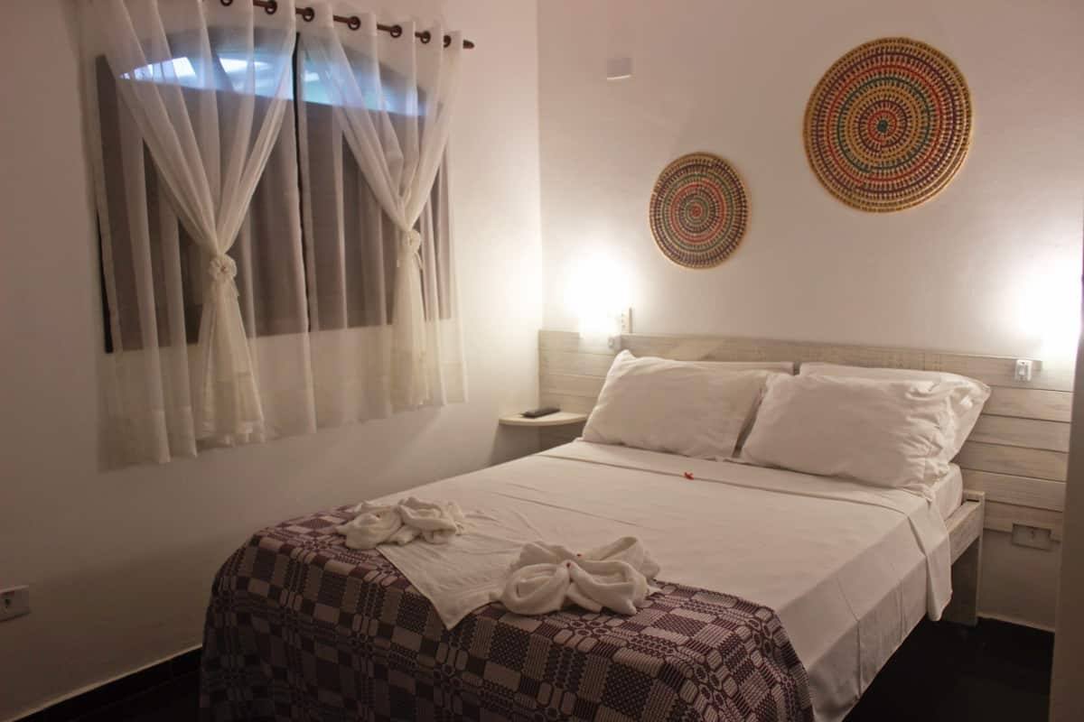 Une chambre d’hôtel comprenant un lit, une table de chevet, une commode, une fenêtre avec des rideaux légers et deux éléments de décoration murale.