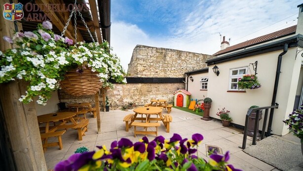 A courtyard with wooden benches and tables, stone walls, and hanging flower baskets. - The Oddfellows Arms (Leeds)