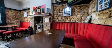 A pub interior with a long wooden table, red upholstered seating, a dartboard, and a stone wall.