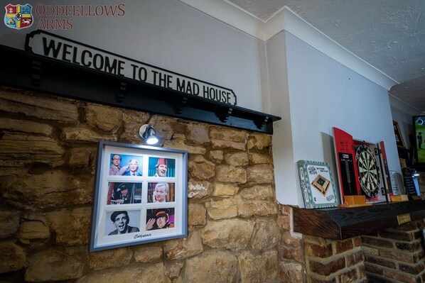 Una pared con un letrero que dice 'Welcome to the Mad House', un collage de fotos enmarcadas y un dardo.