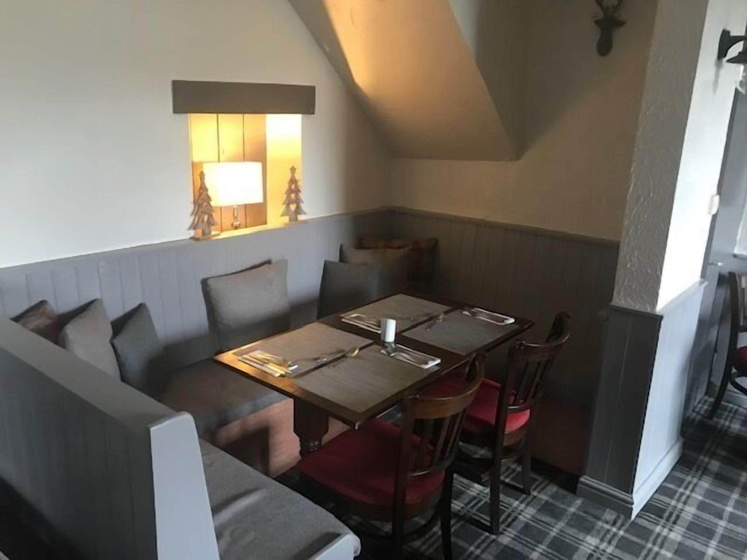 A cozy corner with a wooden table set for two, a cushioned bench, and a small window with a lamp.