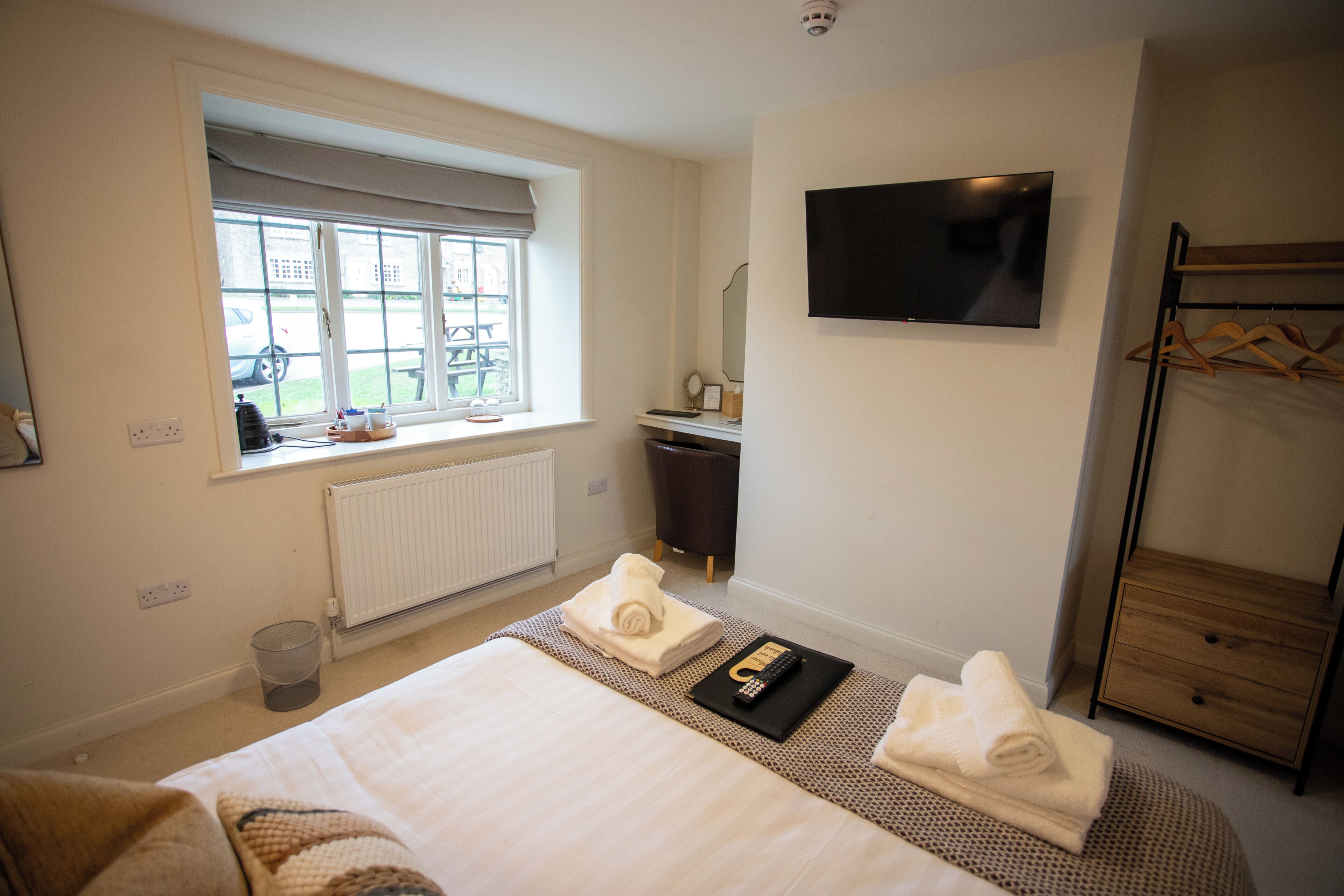 A hotel room with a bed, a TV, a window with a view, a coat rack, and a bedside table with a remote control.