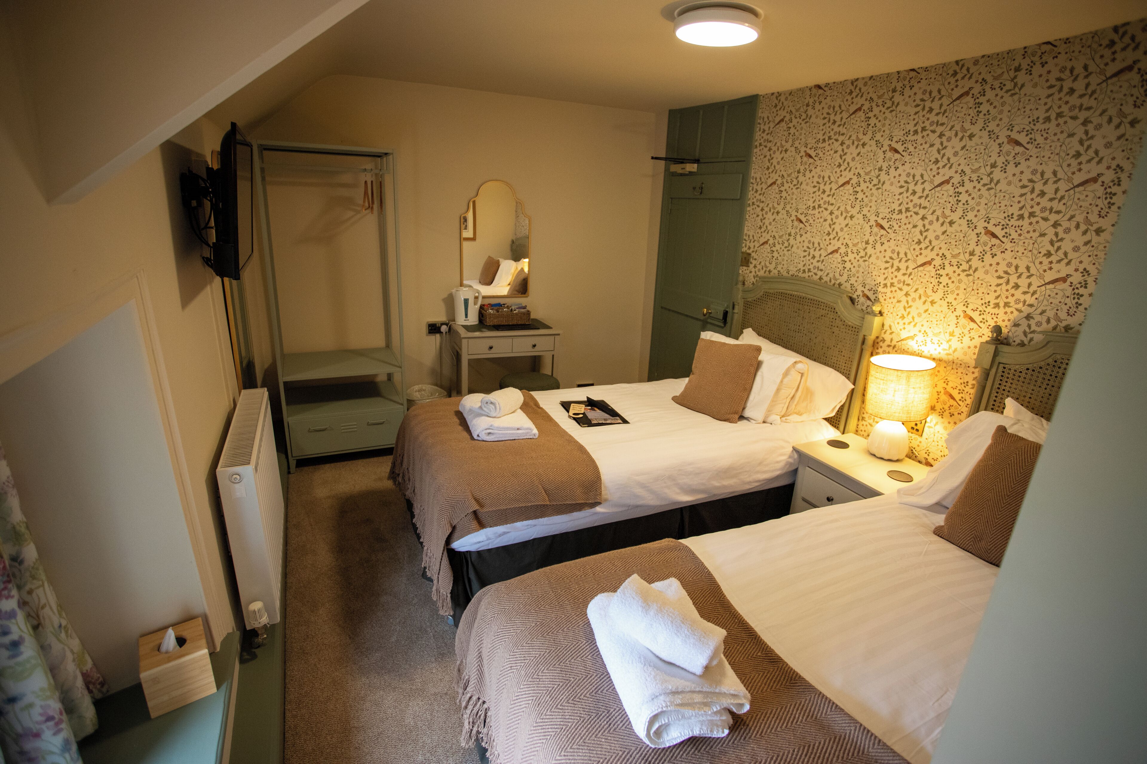 Habitación de hotel con dos camas, una mesita de noche, un espejo, un radiador y una pared con papel tapiz floral.