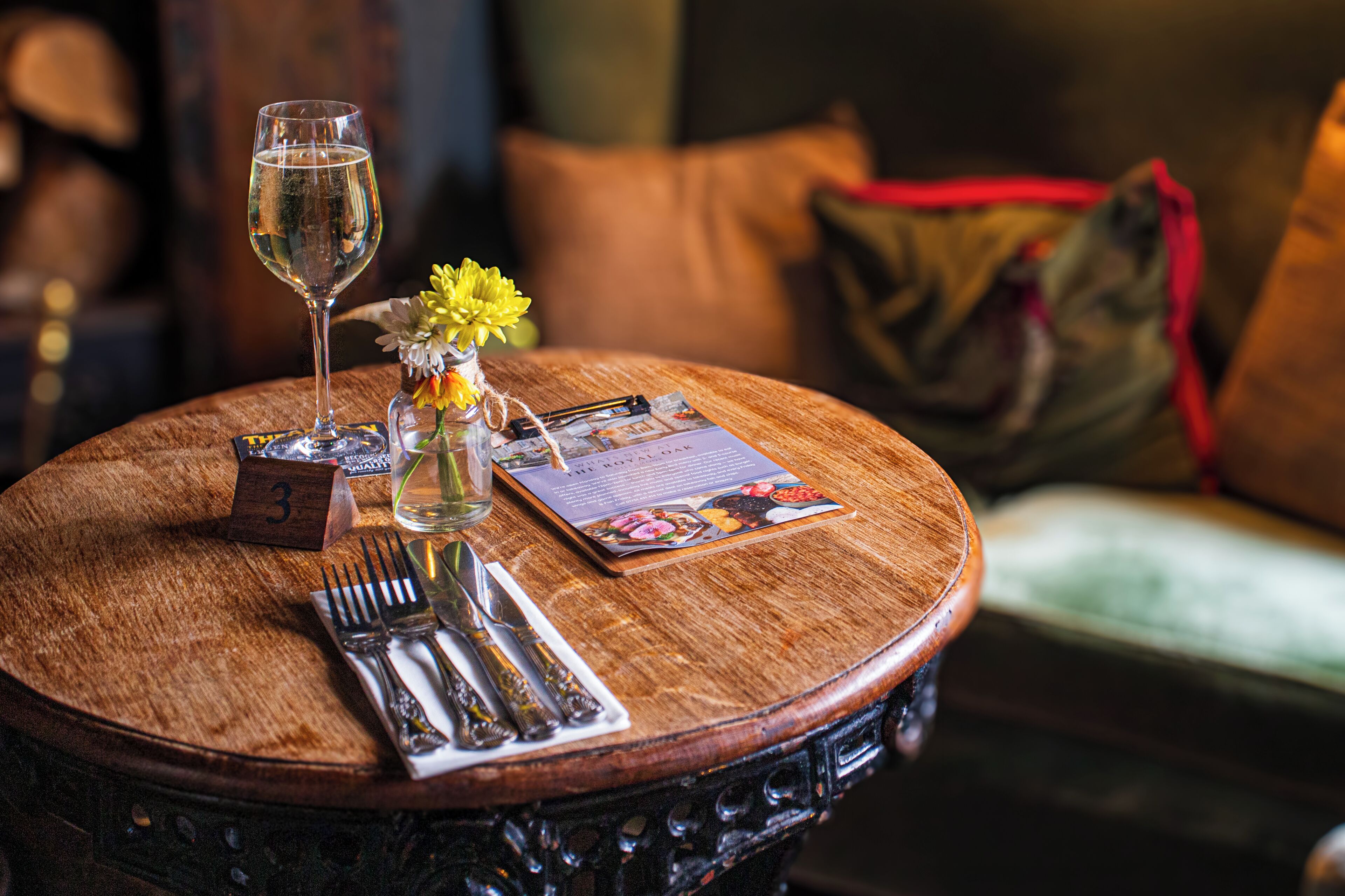 A round wooden table with a menu, a glass of water, a vase with flowers, and a number three sign.
