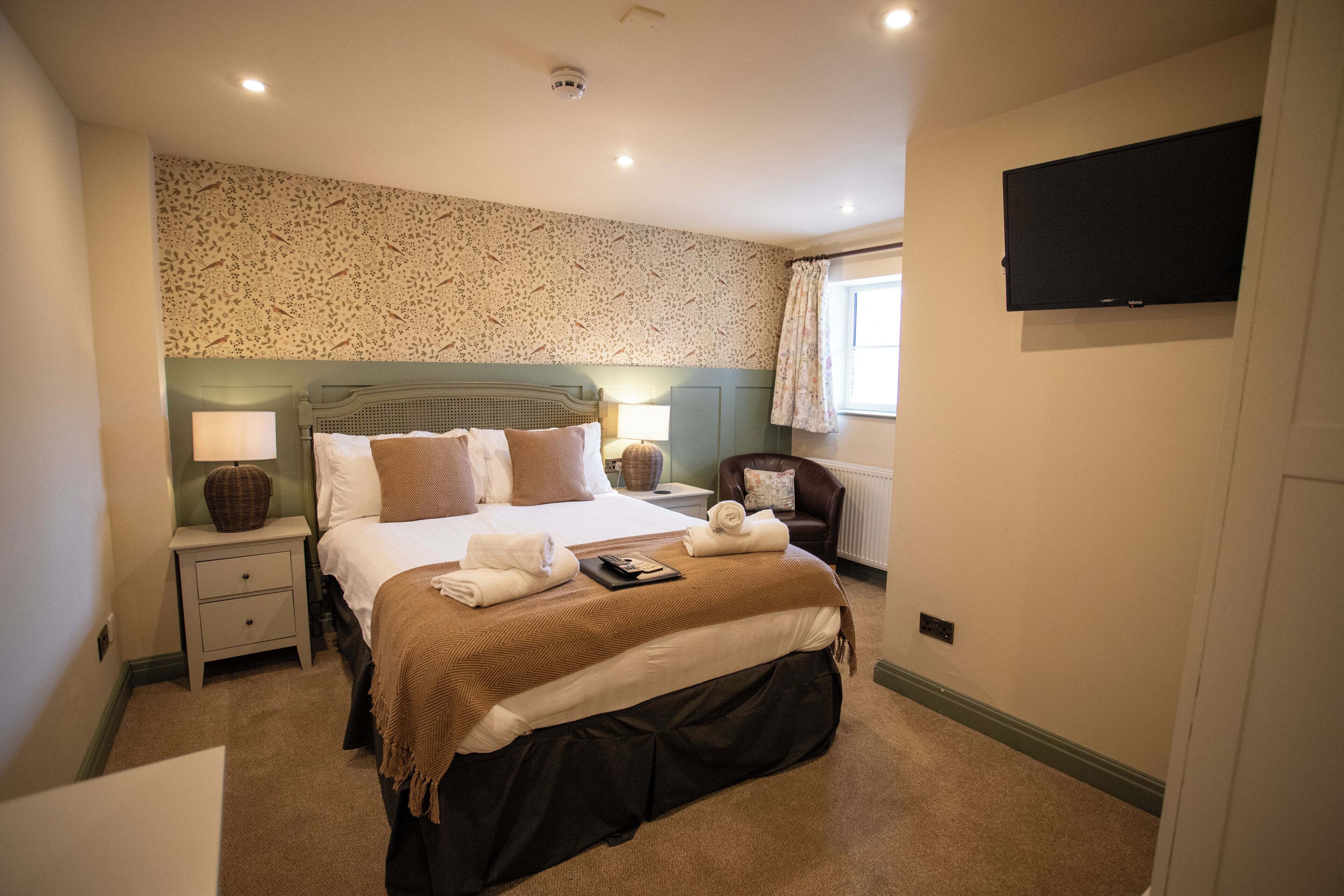 Habitación de hotel con cama, mesitas de noche, lámpara, silla, televisor y ventana con cortinas.