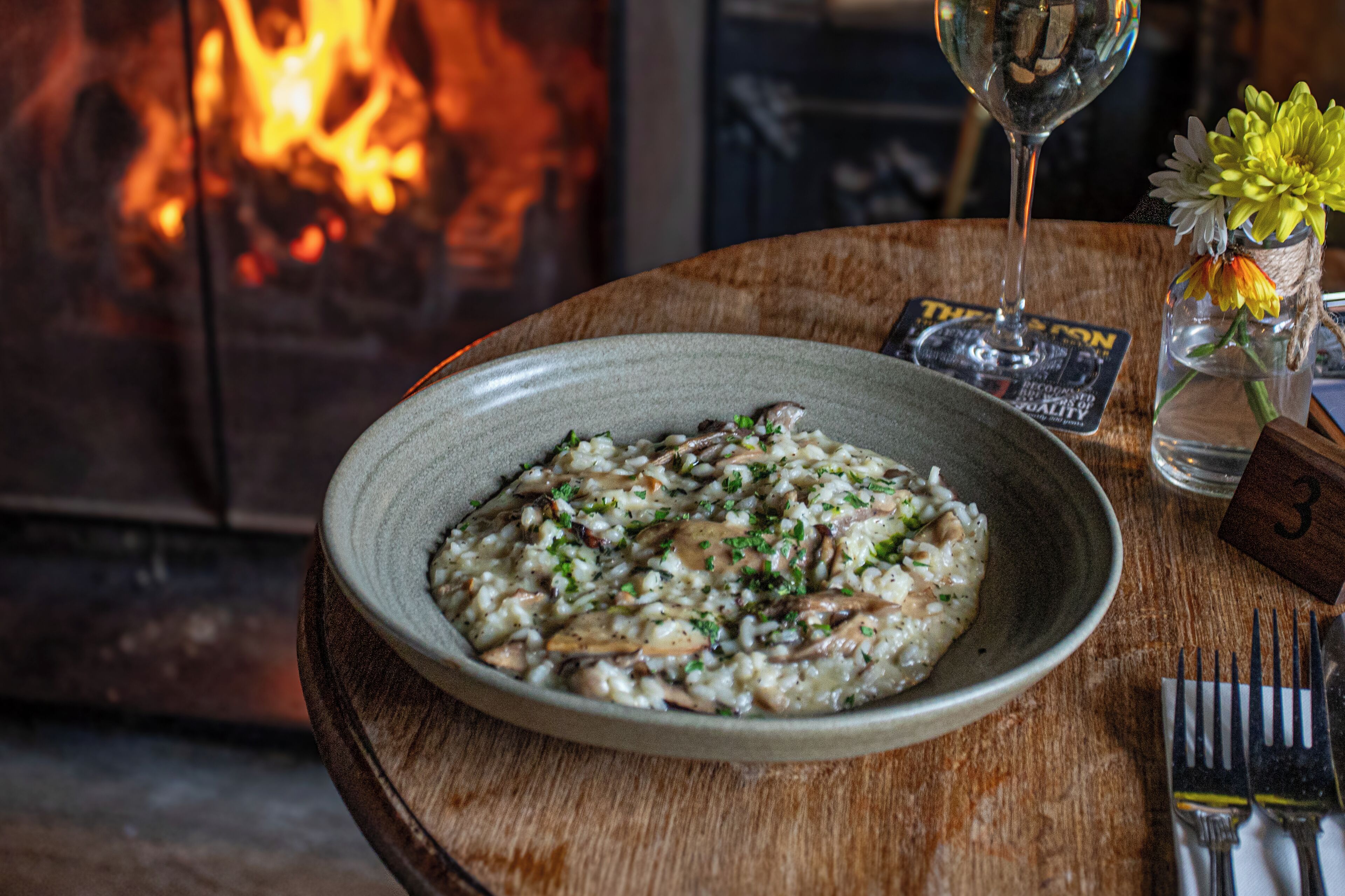A plate of risotto with mushrooms and herbs, a glass of wine, a vase with flowers, and a fireplace in the background.