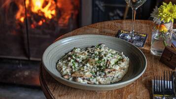 A plate of risotto with mushrooms and herbs, a glass of wine, a vase with flowers, and a fireplace in the background.