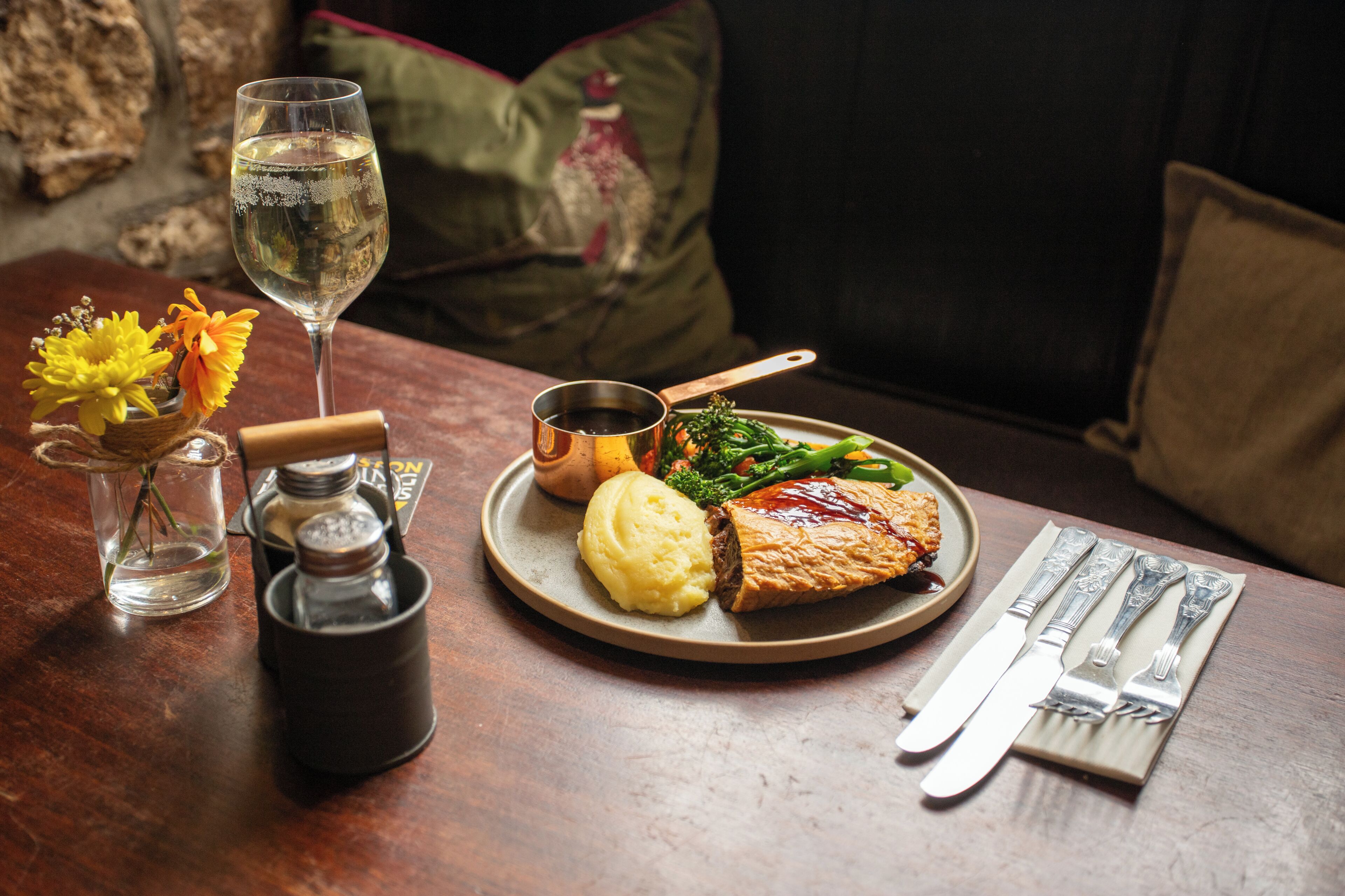 A dining table with a plate of food, a glass of wine, salt and pepper shakers, and a knife and fork set.