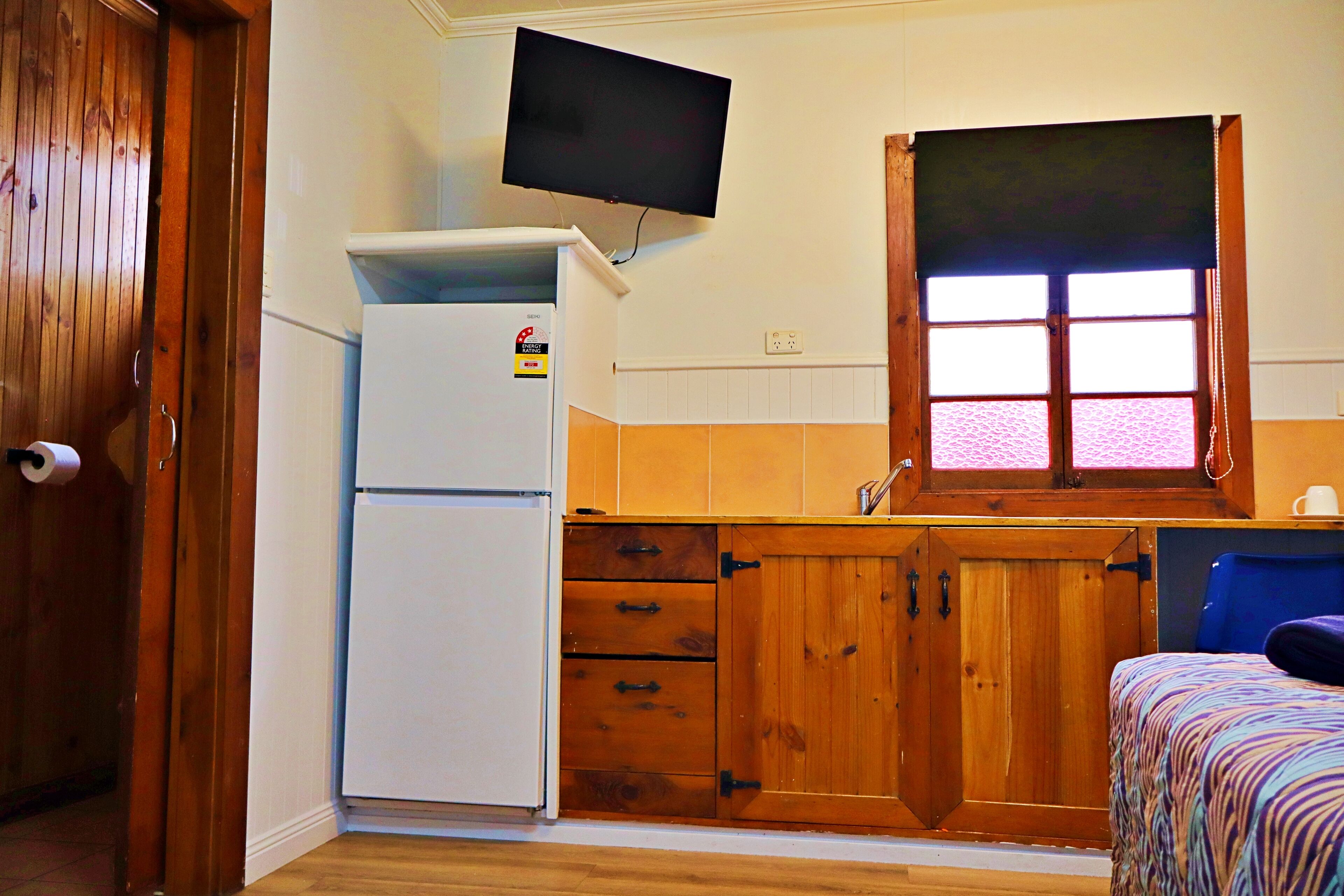 A compact kitchen with a refrigerator, wooden cabinets, a window with blinds, and a television mounted above the counter.