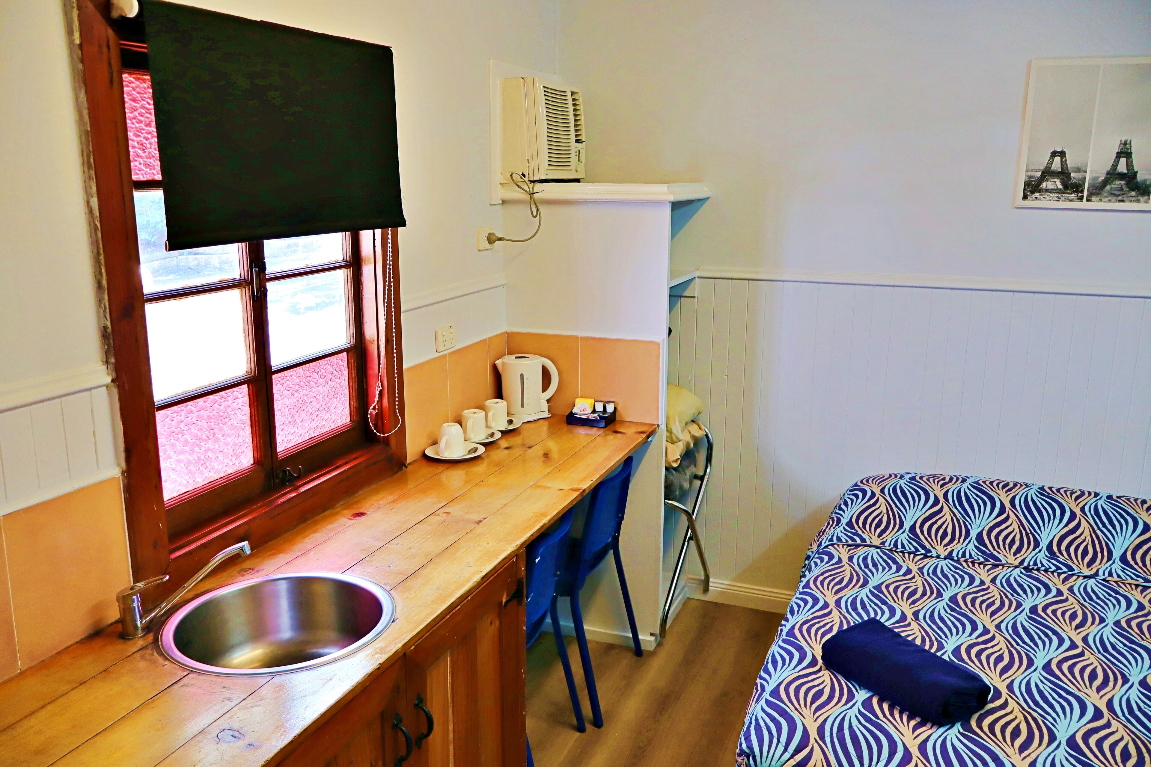 Una cocina compacta con un lavamanos, una ventana con persianas, un hervidor y una silla.