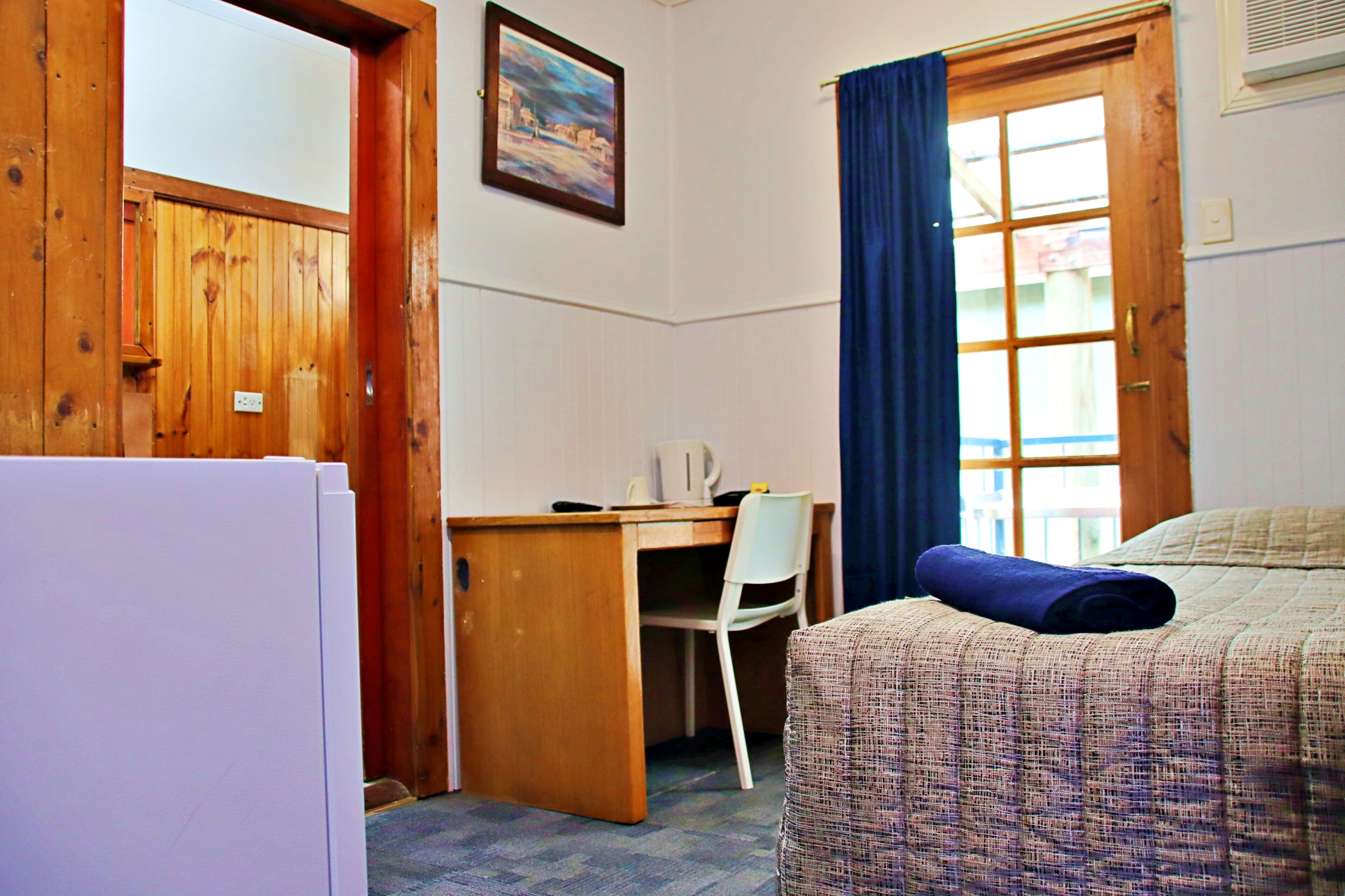 Una habitación con escritorio de madera, una silla, una cama con colcha estampada y una ventana con cortinas azules.