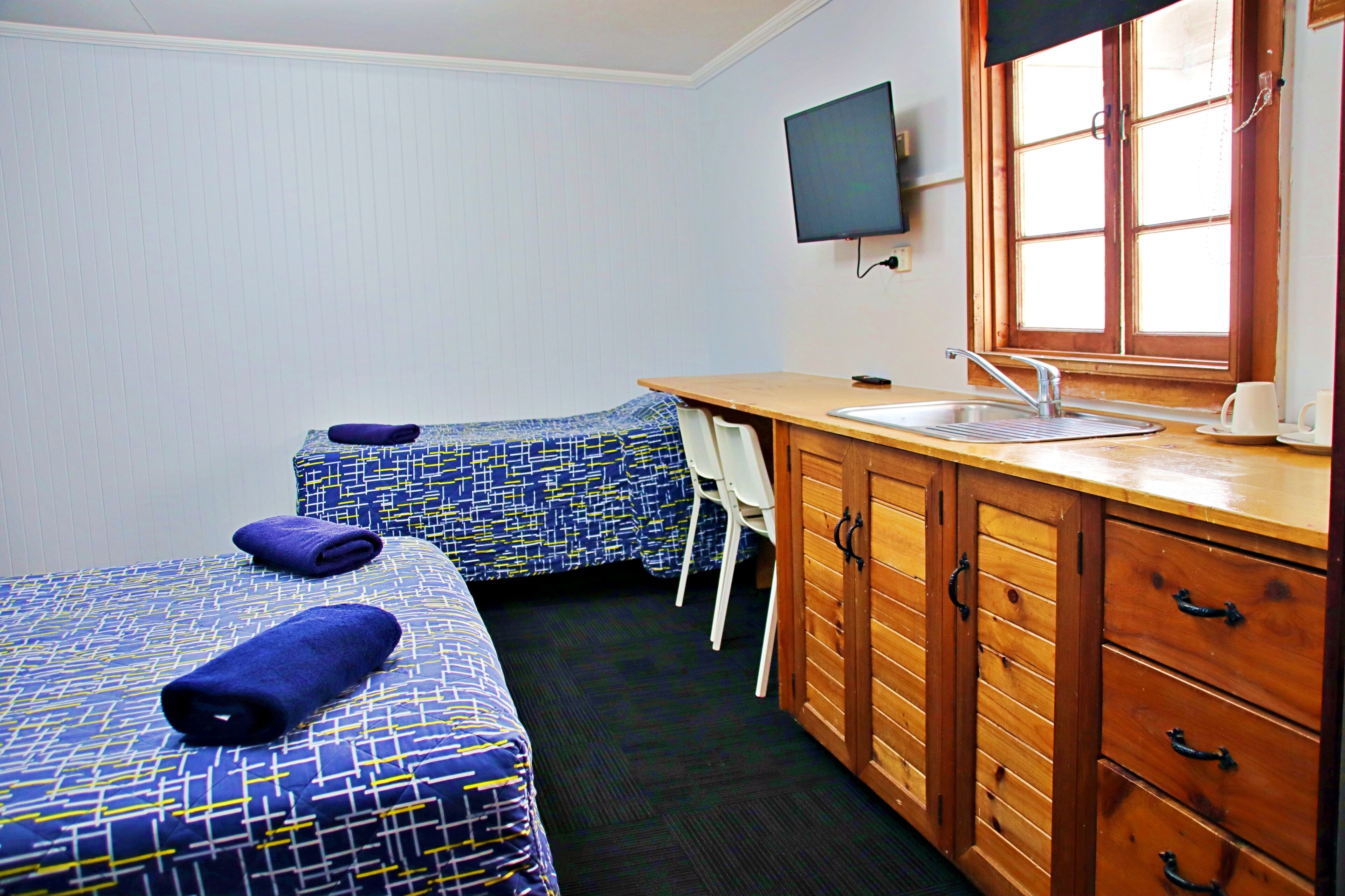 A small, clean room with two beds, a wooden cabinet with drawers, a sink, and a window with blinds.