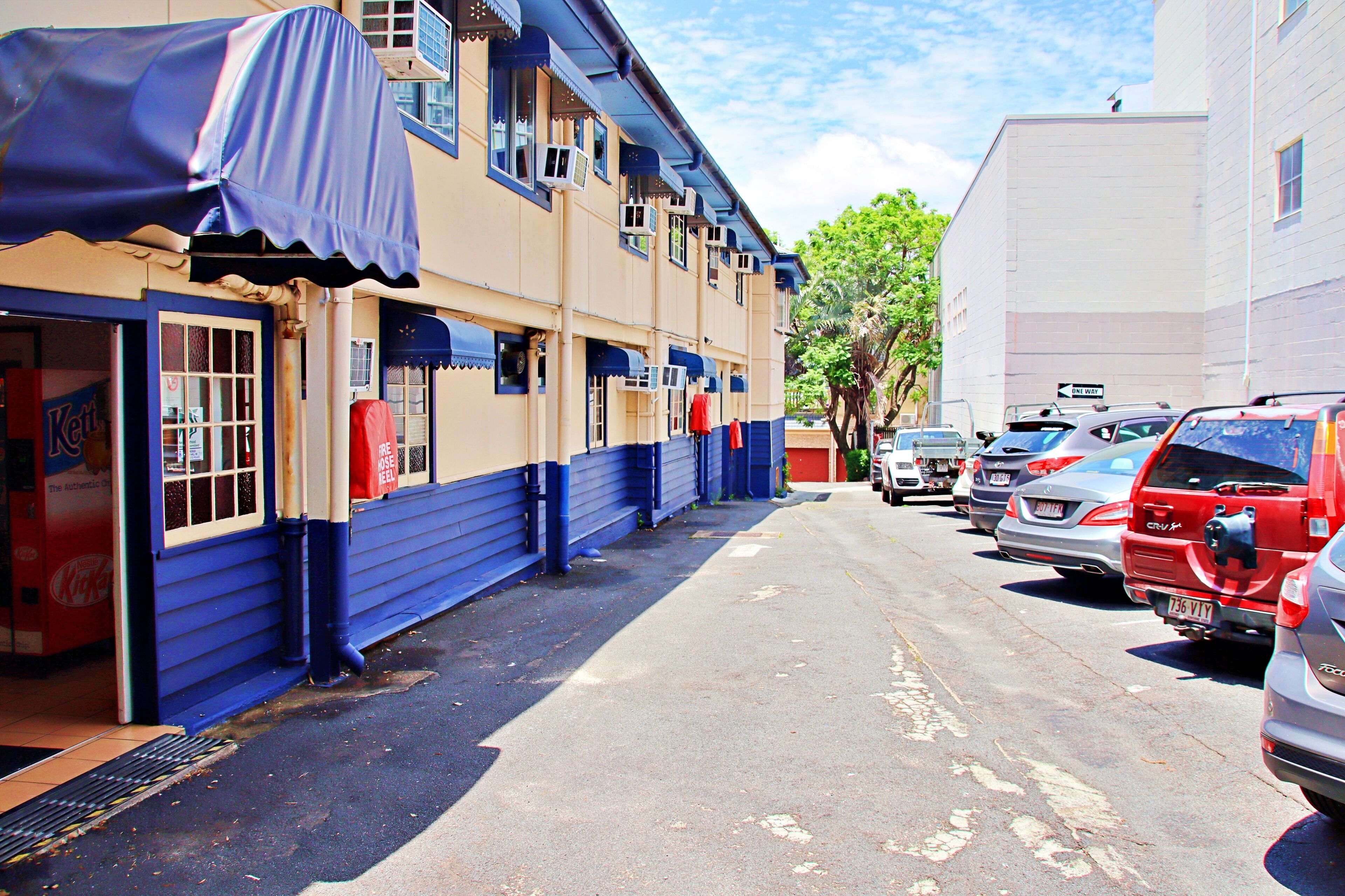 Una fila de coches estacionados a lo largo de una calle, con un edificio de paredes azules y beiges y un toldo cubierto.