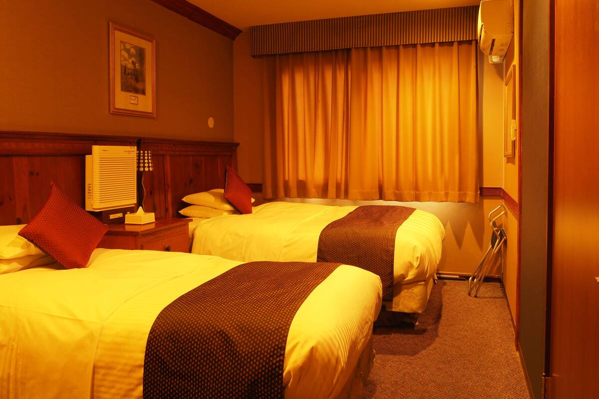 A hotel room with two beds, a ceiling fan, a window with curtains, a bedside table with a lamp, and a wall-mounted air conditioning unit.