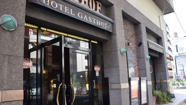 The exterior of Hotel Gasthof with a prominent entrance, a signboard, and a small outdoor seating area with plants. - Hotel Gasthof (Kagoshima)