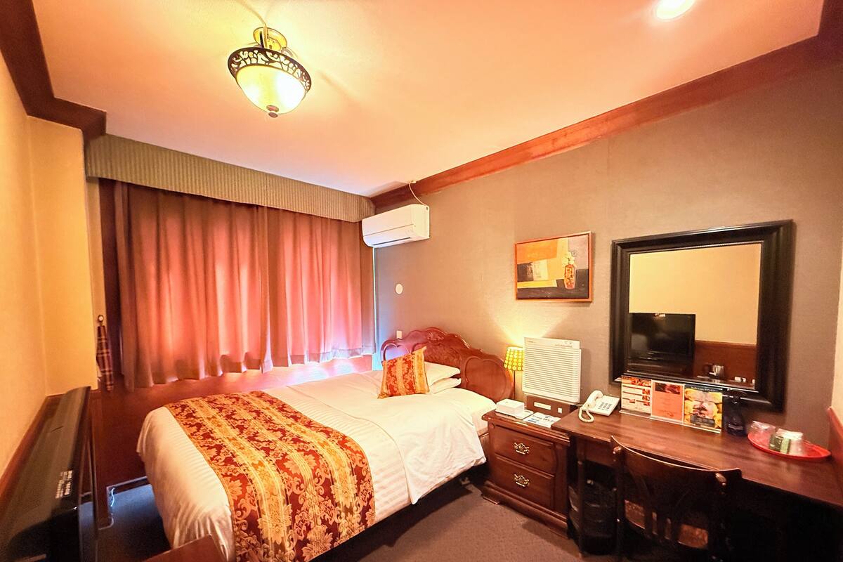 A hotel room with a bed, a desk, a chair, a telephone, a mirror, and a window with curtains.