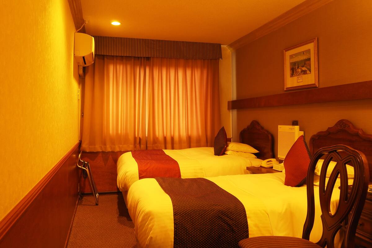 A hotel room with two beds, a wooden headboard, a small table, a chair, a window with curtains, and a picture on the wall.