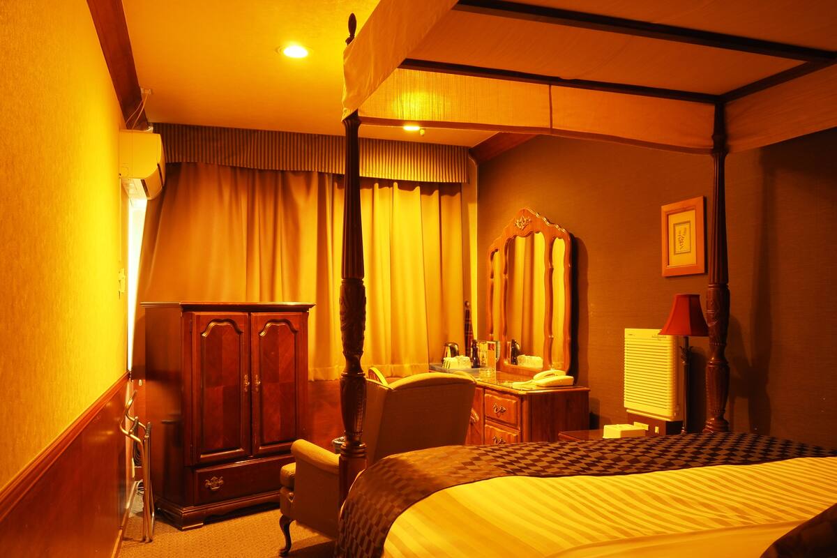 A hotel room with a four-poster bed, a wooden wardrobe, a desk with a chair, a mirror, and a bedside lamp.