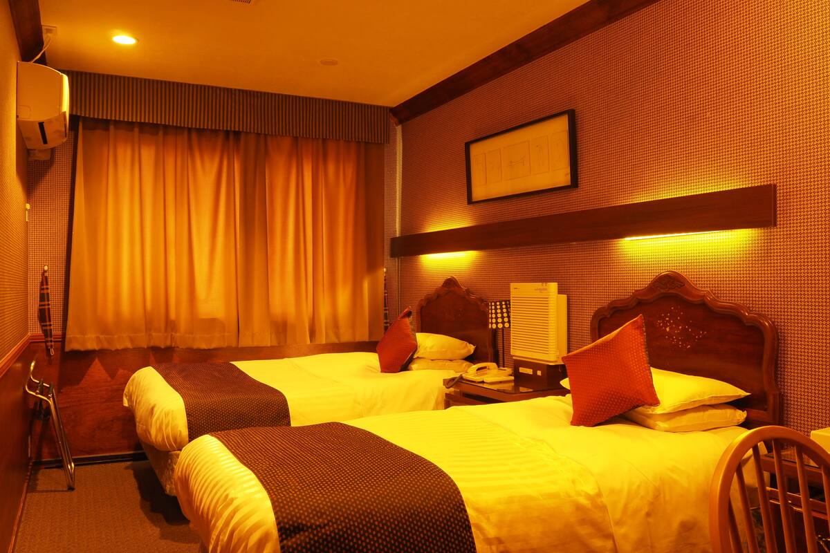 A hotel room with two beds, a bedside table with a telephone, a wall-mounted air conditioner, and a framed picture on the wall.