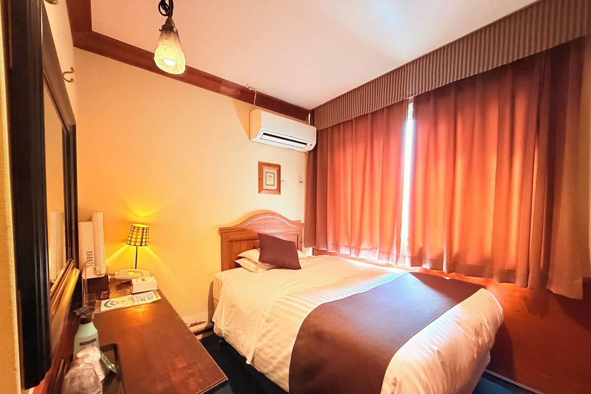 A hotel room with a bed, bedside table, lamp, air conditioning unit, and a window with curtains.