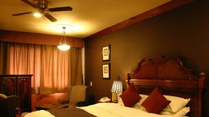 A hotel room with a bed, a desk, a chair, a ceiling fan, and a nightstand with a lamp. - Hotel Gasthof (Kagoshima)