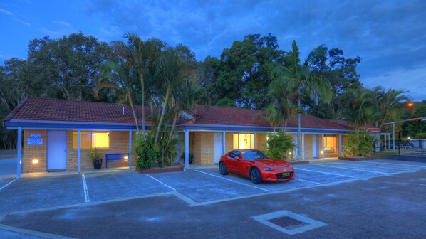 Un motel con un coche rojo estacionado en el estacionamiento, rodeado de palmeras y un cielo despejado.