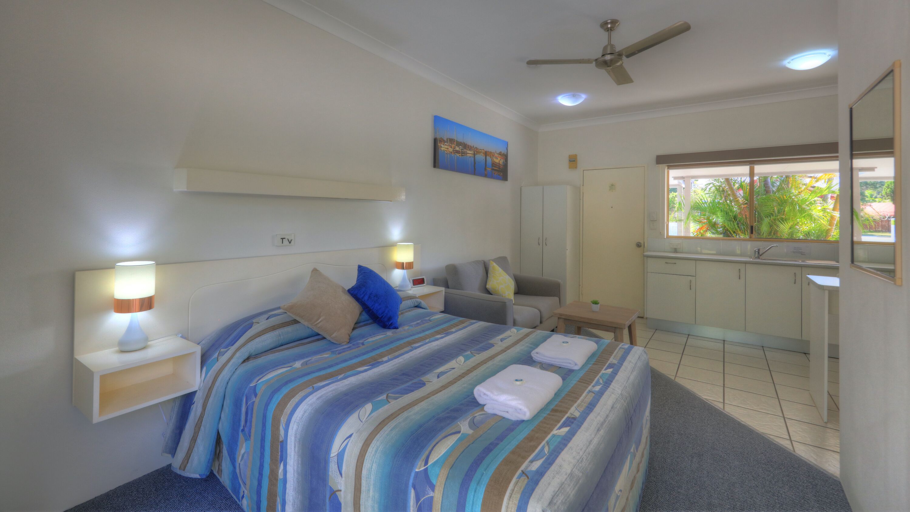 A hotel room with a bed, bedside lamps, a TV, a painting, a ceiling fan, a window with a view of greenery, and a small table with a plant.