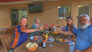 A group of people sitting around a wooden table with food and drinks, in a room with brick walls and a TV mounted on the wall.