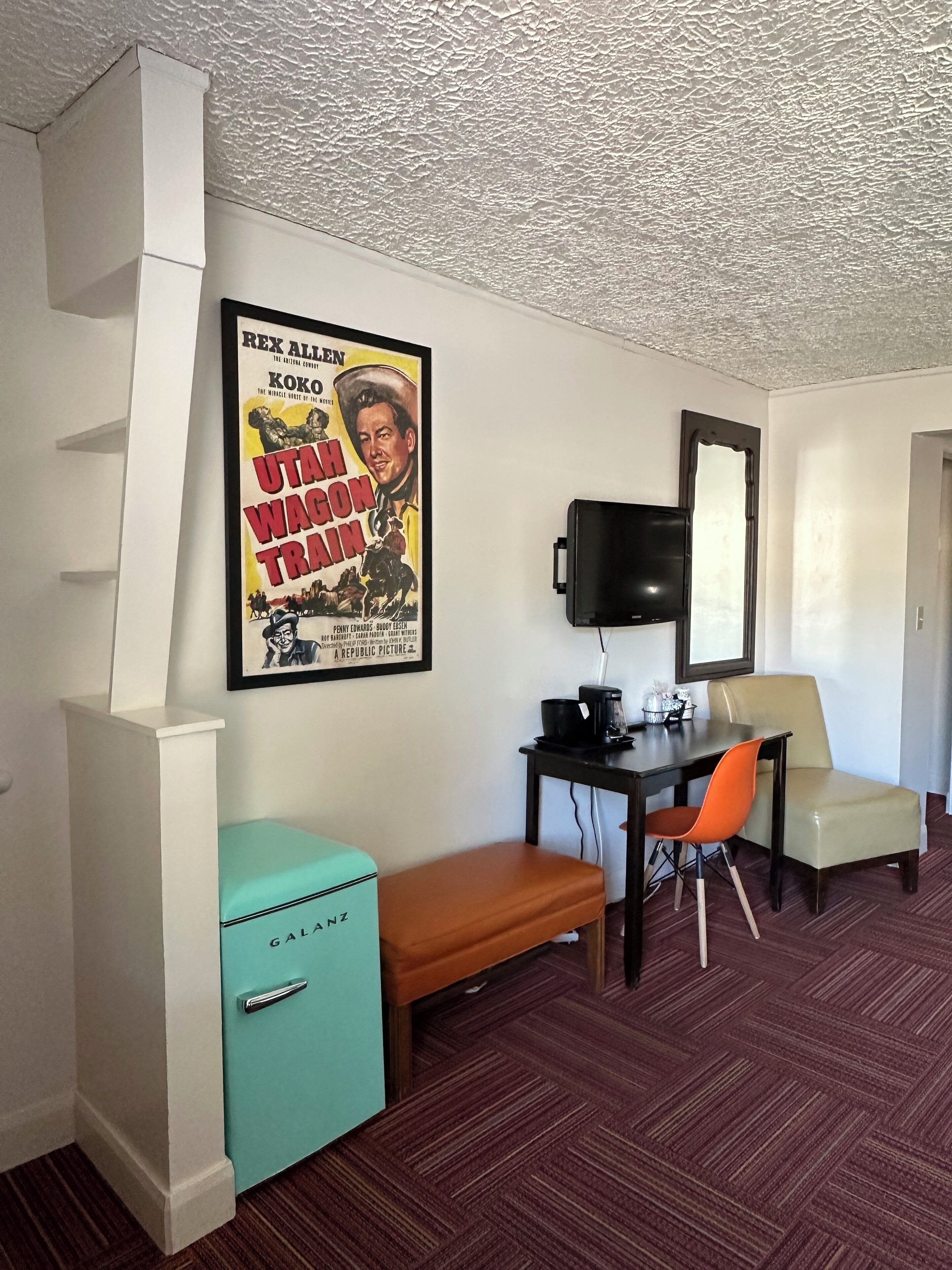 A hotel room with a teal minibar, a wooden bench, a dining table with a chair, a flat-screen TV, and a poster of 'Utah Wagon Train'.