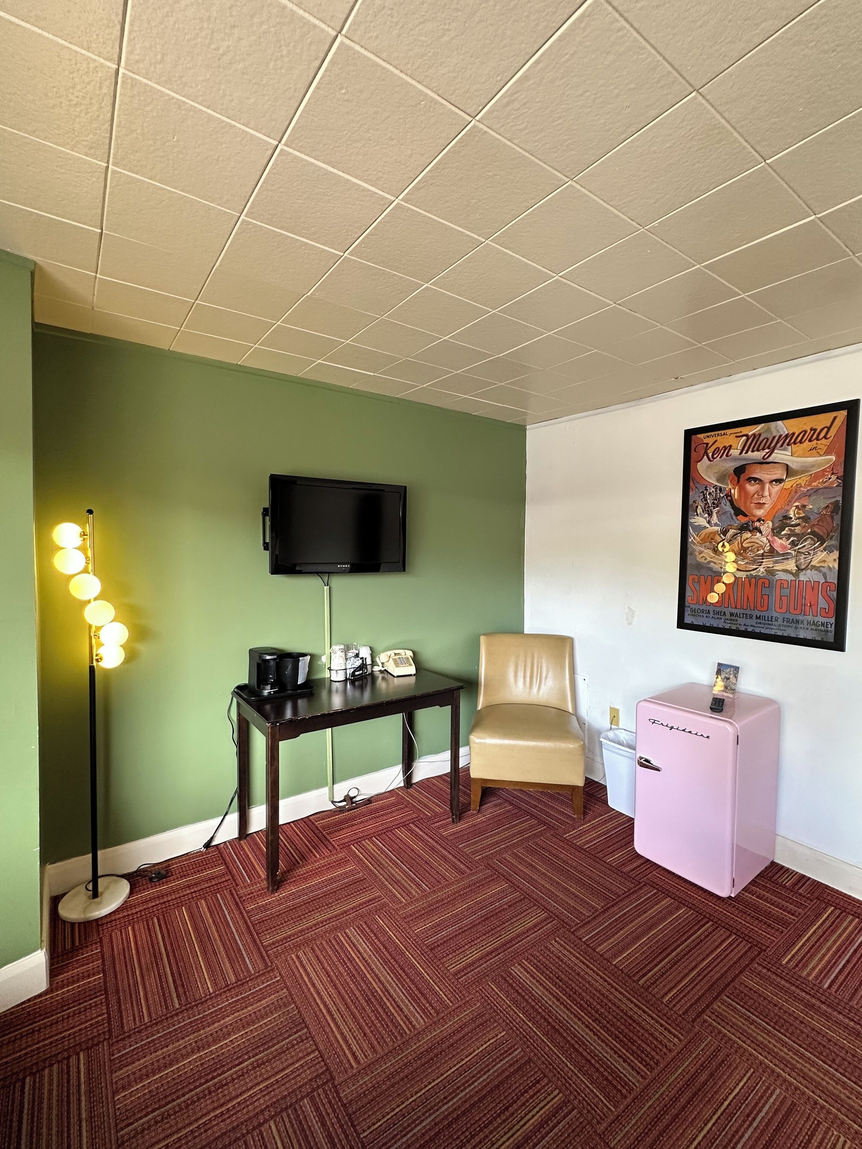 A room with a green wall, a flat-screen TV mounted on the wall, a small table with a telephone and a coffee maker, a chair, a pink refrigerator, and a poster on the wall.