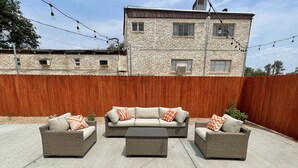 Een terras met drie rieten banken en een salontafel, een houten schutting en een stenen gebouw op de achtergrond.