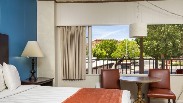 Een hotelkamer met een bed, een tafel, twee stoelen, een lamp en uitzicht op een stadsbeeld en bomen.