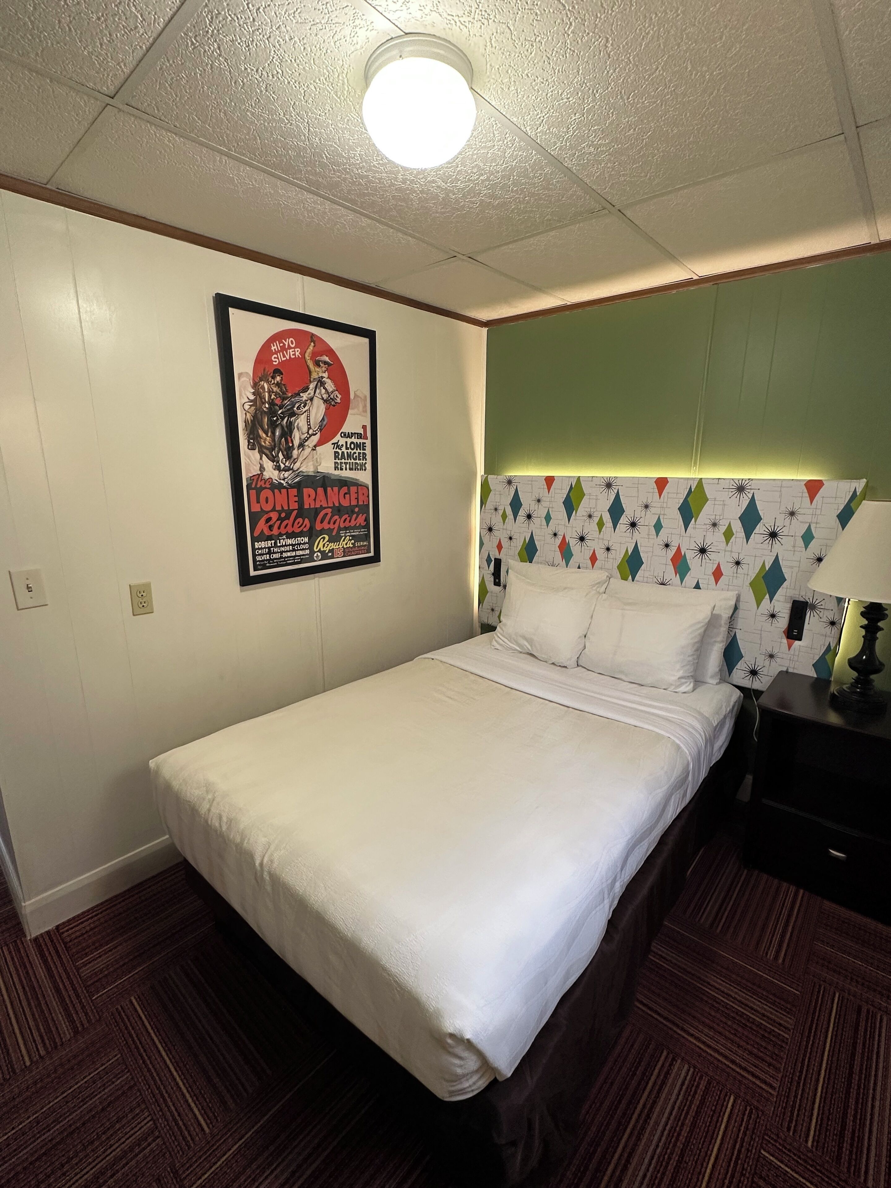 Habitación de hotel con una cama individual, cabecera con diseño geométrico, un cartel en la pared y una lámpara en el techo.
