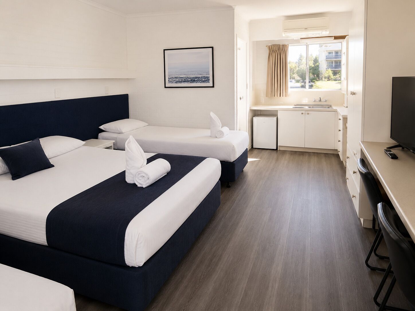 A hotel room with two beds, a small kitchenette, a television, and a window with curtains.