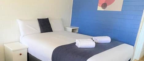 Una cama bien hecha con sábanas blancas y un edredón gris oscuro, una mesita de noche blanca con un cajón, y una pared azul con un cuadro abstracto geométrico.