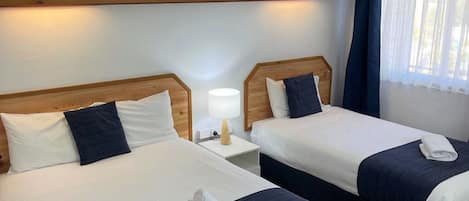 Een hotelkamer met twee bedden, elk met wit en marineblauwe beddengoed, een nachtkastje met een lamp en een raam met gordijnen.