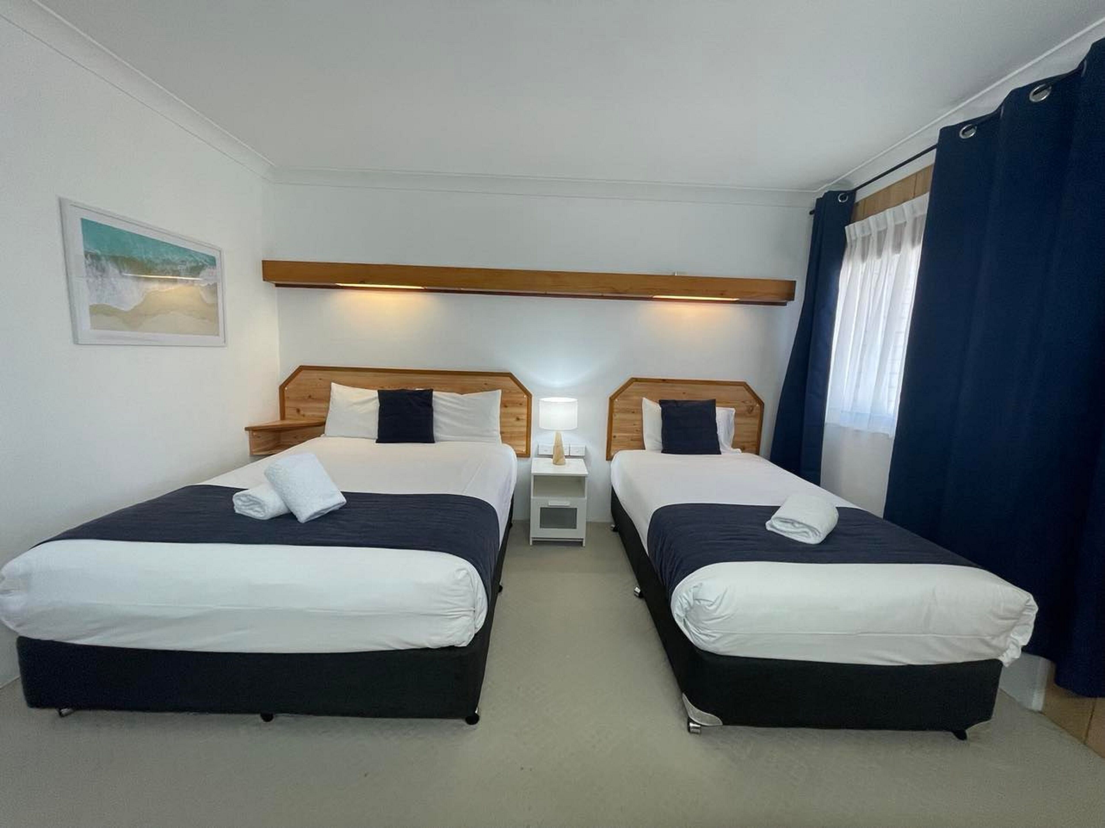 A hotel room with two single beds, a nightstand between them, a lamp, and a painting on the wall.