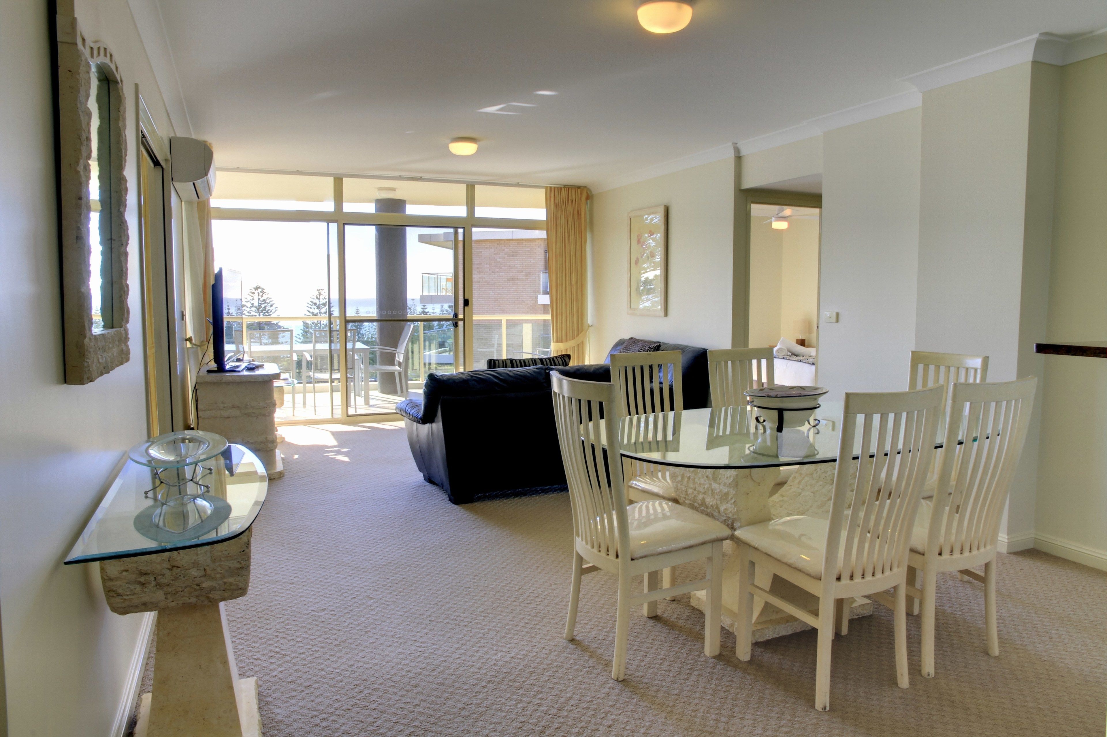 A dining area with a glass table and white chairs, a black sofa, and a view of the outside balcony.