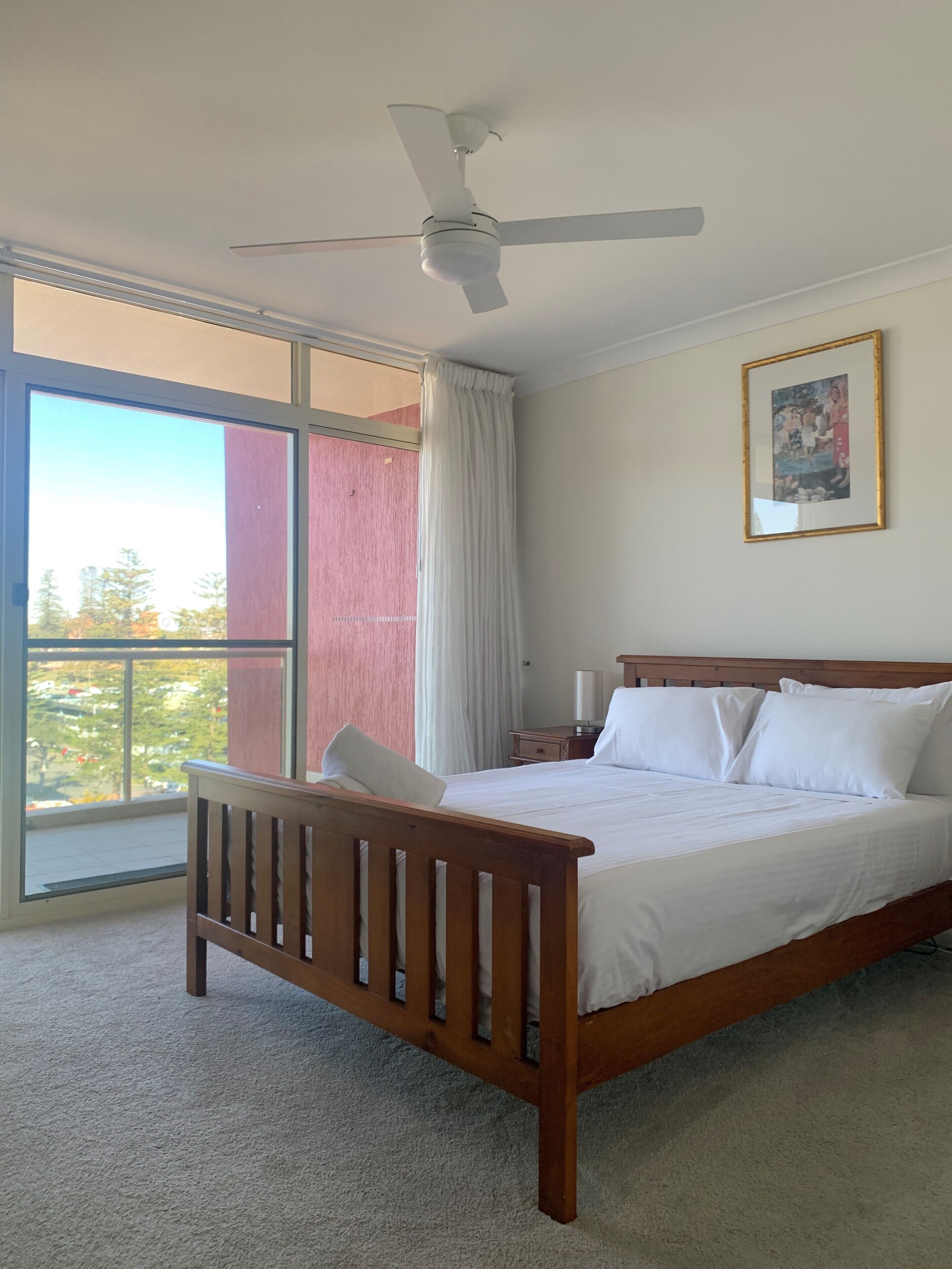 Un dormitorio con cama, mesita de noche, ventilador de techo, ventana con cortinas e imagen enmarcada en la pared.