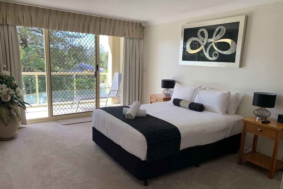 Habitación de hotel con una cama grande, una mesita de noche con lámpara, vistas al exterior a través de una puerta corredera y una planta en maceta.