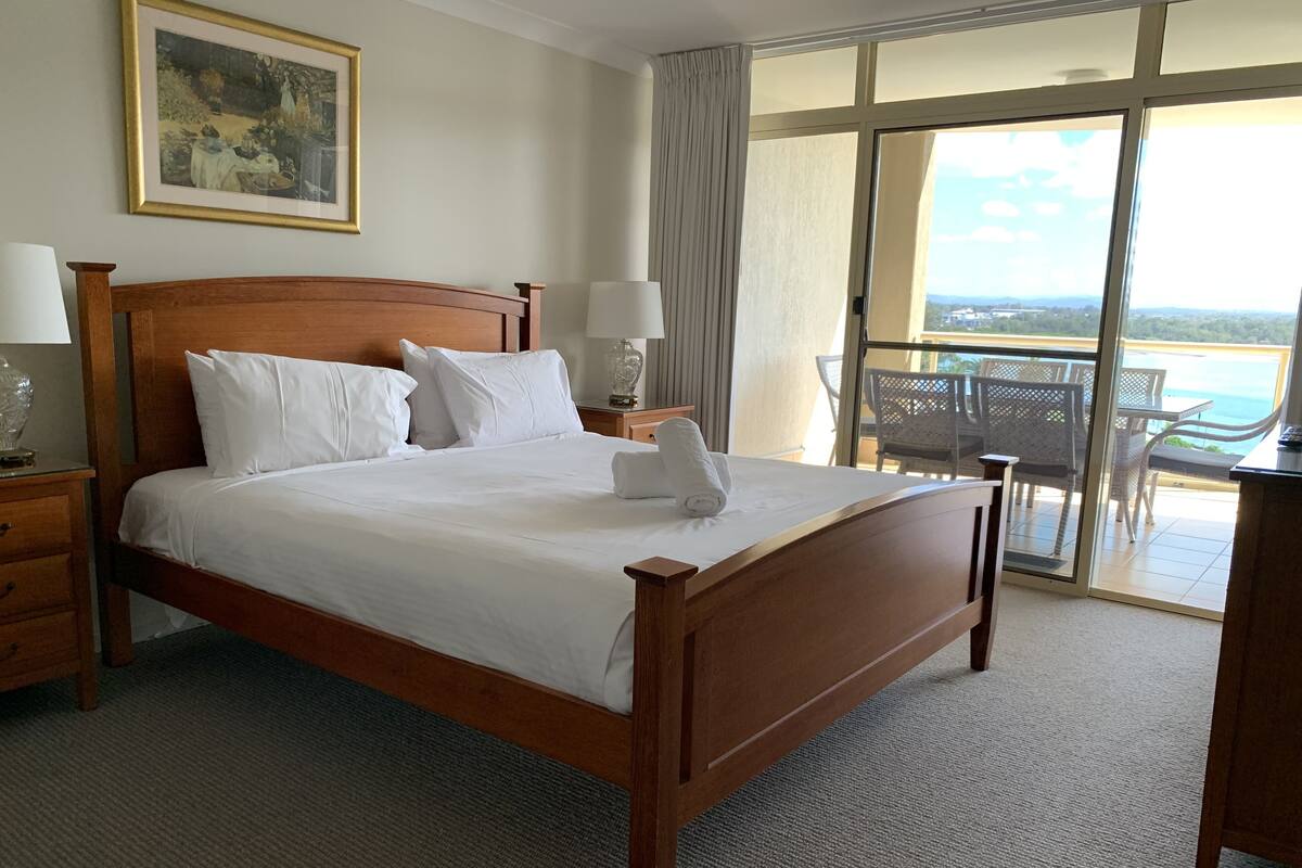 Habitación de hotel con cama de madera, ropa de cama blanca, un cuadro en la pared, balcón con mobiliario exterior y vista al mar.
