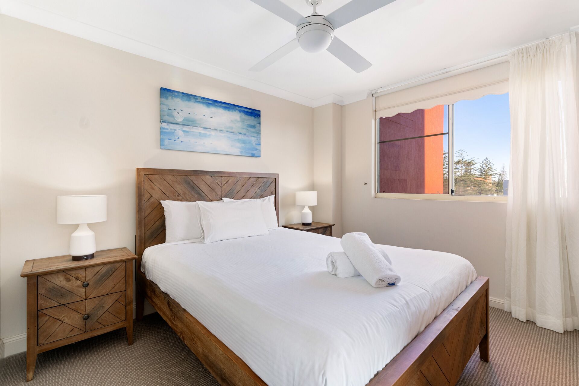 A hotel room with a large bed, wooden headboard, two bedside tables with lamps, a ceiling fan, a window with curtains, and a painting on the wall.